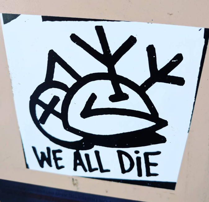 A sticker that reads “we all die,” photographed.